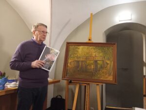 Jurij Wuschansky steht neben einem Gemälde eines Holzstegs über einem Bach in einem herbstlichen Wald, hält ein Foto in der Hand, trägt einen lila Pullover und eine Hose.