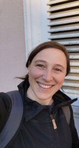 Heidi Pinkepank trägt eine schwarze Jacke und einen Rucksack, steht vor einer weißen Wand mit Lüftungsgitter