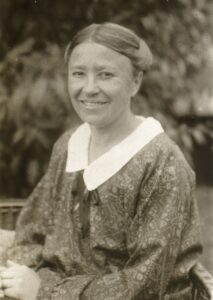 Sitzende Frau mit gemustertem Kleid und weißem Kragen vor unscharfem Laubhintergrund.